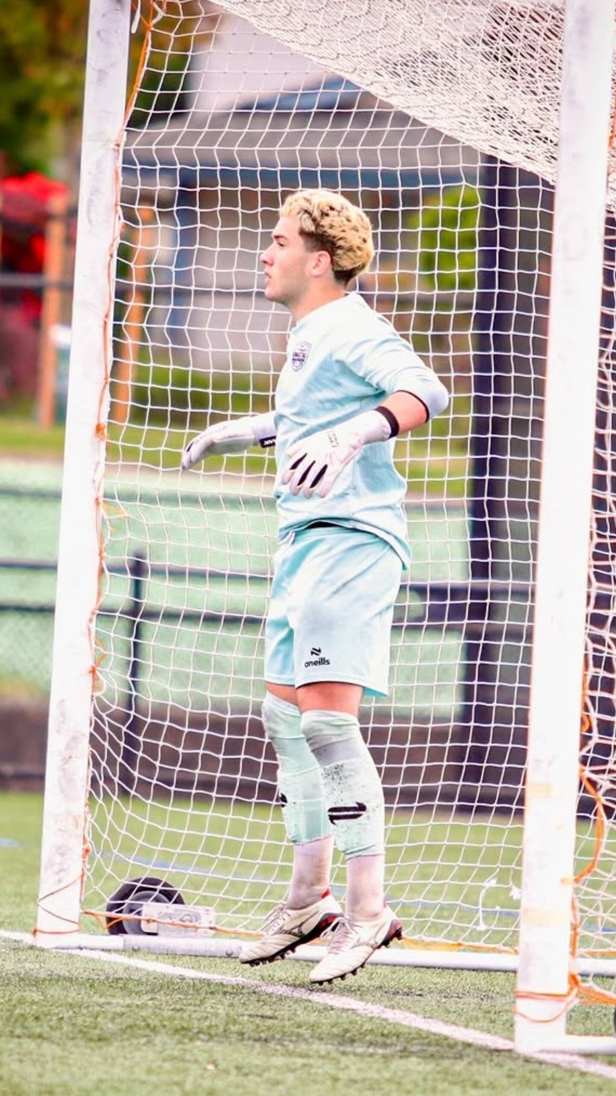 A soccer goalkeeper in a light blue and white uniform stands in front of a white goal net on a green turf field, wearing RANK Goalkeeper gloves and looking upward.