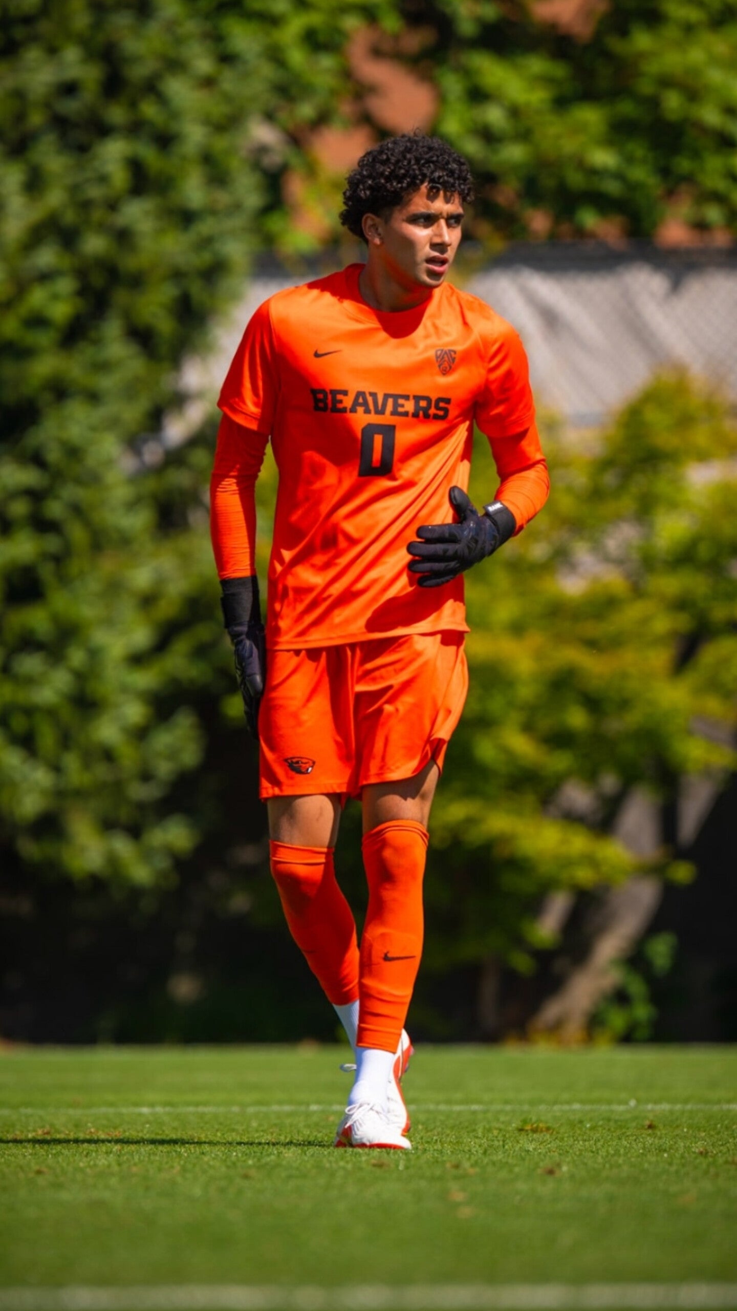 A soccer player wearing an orange long-sleeve jersey, shorts, and socks with black gloves, walking on a green grass field with trees in the background. The jersey has 'BEAVERS' and the number '0' printed on the front.