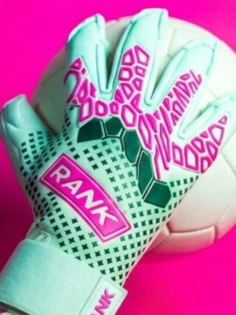 From the elite, top quality, emerging and goalkeeper favorite, the new glove from RANK Goalkeeping. A close-up of a goalkeeper glove with a pale mint green and white patterned palm, bright pink grip text, and green hexagonal accents, resting on a white soccer ball against a vivid pink background. The brand name RANK Goalkeeping is visible on the wrist strap.