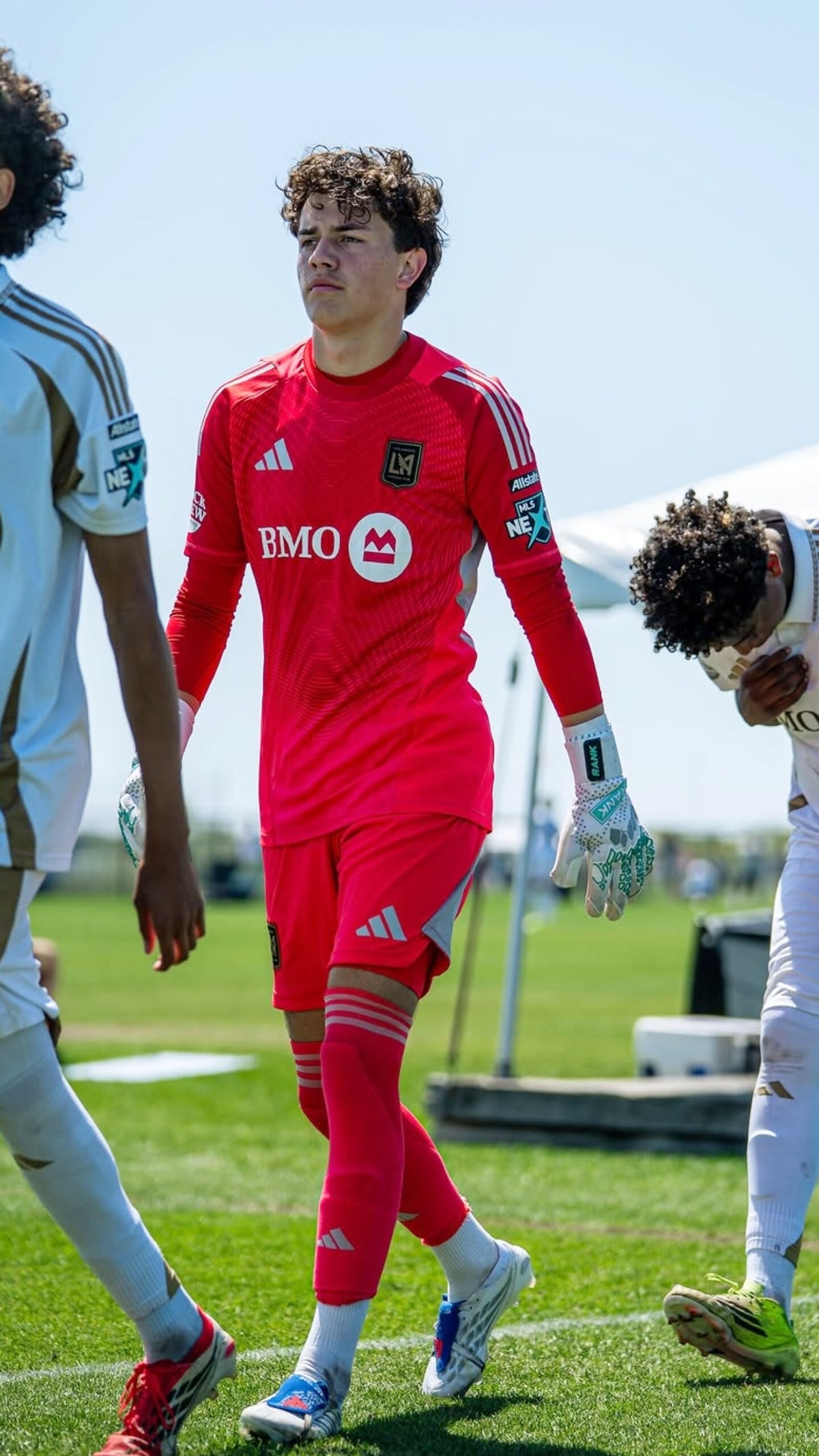 A soccer player wearing goalkeeper jersey, RANK Goalkeeping gloves and matching shorts walks on a green grass field under a blue sky, with teammates partially visible on both sides.
