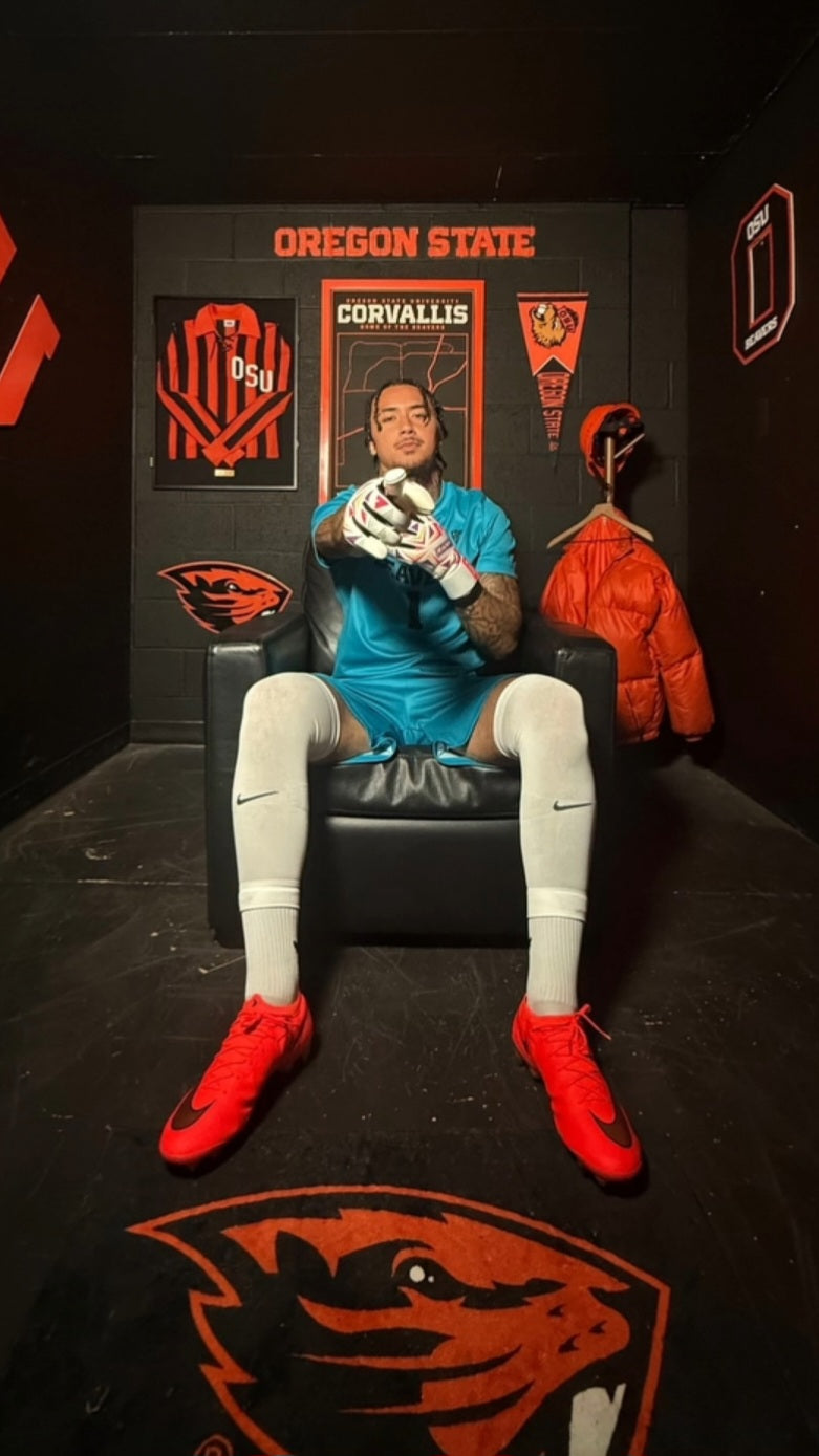 Goalkeeper John Nicholson, Oregon State University, sits in a black leather chair inside a themed Oregon State locker room. The person is wearing a bright blue jersey, white socks, and red cleats, with Oregon State branding and a beaver logo on the floor behind them.