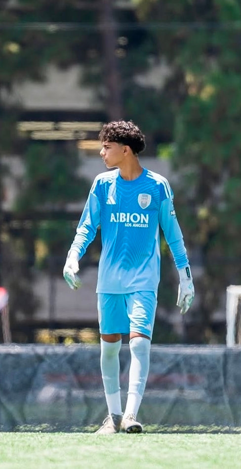 A soccer player wearing a light blue long-sleeve jersey and matching shorts with white socks and gloves, standing on a grassy field with trees and a fence in the background.