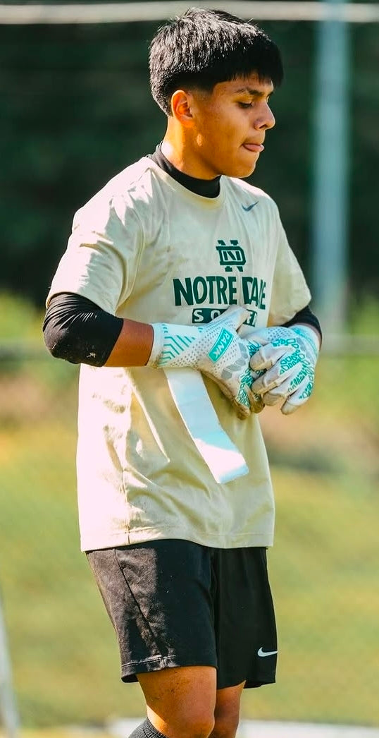 A young soccer player wearing a Notre Dame Saints beige practice jersey, black shorts, and white goalkeeper gloves standing on a grassy field.