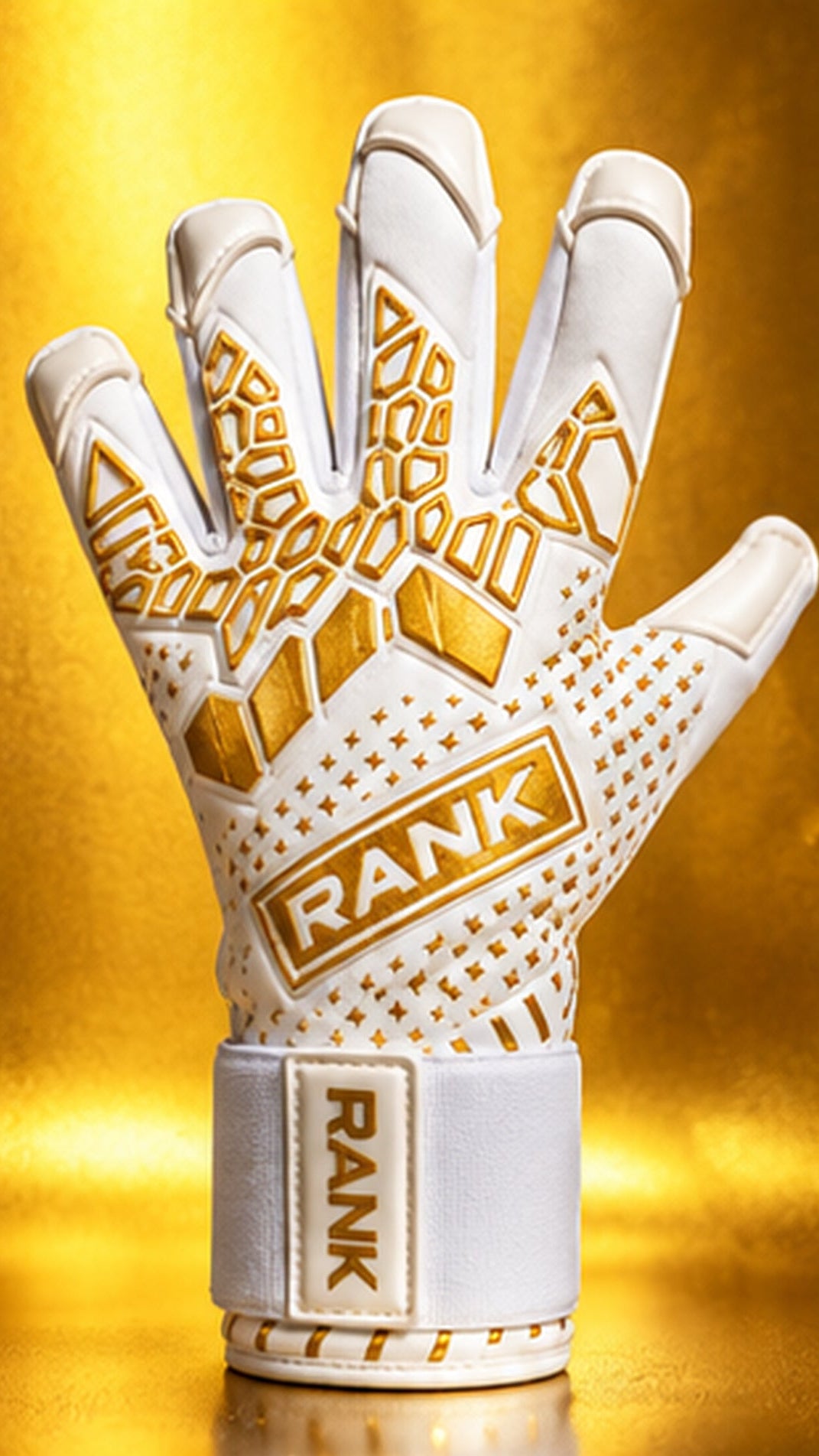 A white and gold goalkeeper glove by RANK Goalkeeping with gold accents and the brand name 'RANK' printed on the back and wrist strap, set against a reflective gold background.