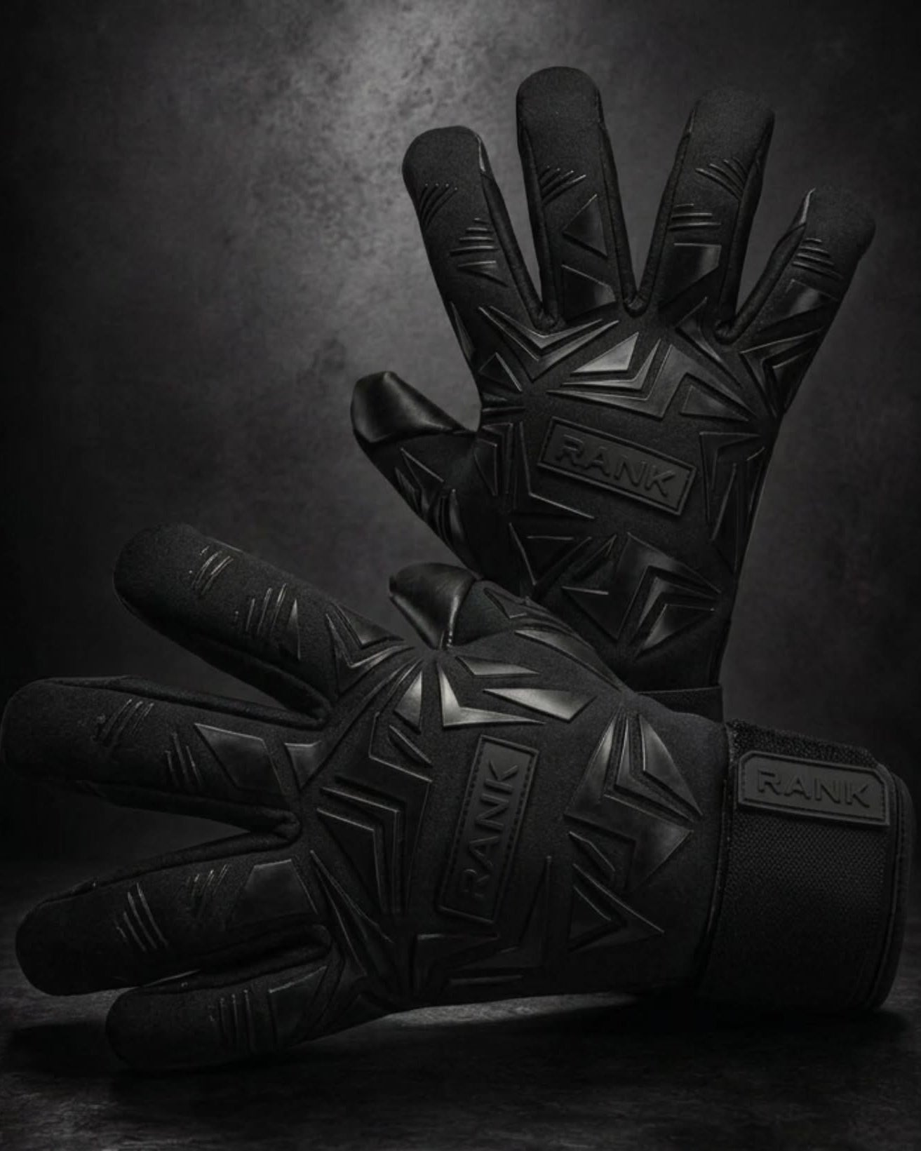 A pair of black protective gloves with geometric embossed patterns and visible RANK branding, displayed against a dark textured studio background.